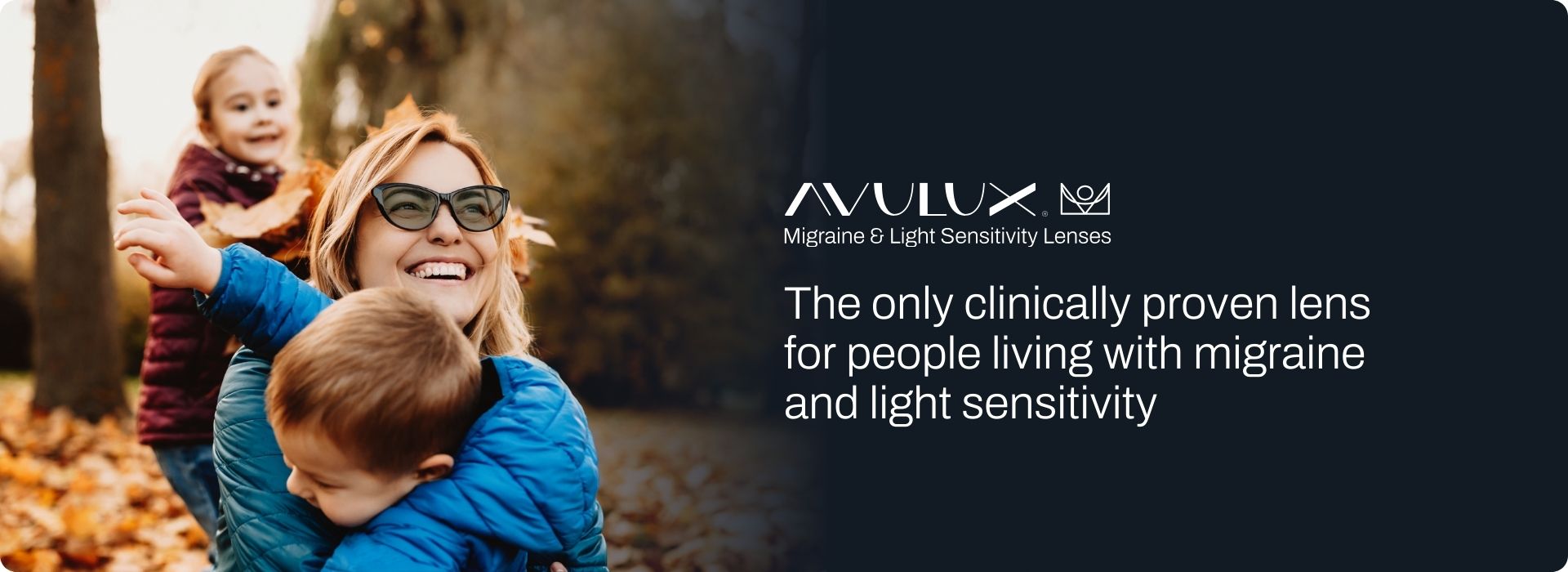 A blonde woman is wearing Avulux migraine lenses and smiling. Her two blonde children under the age of 6 are holding on to her. Everyone is smiling and the background is being outside in the sunlight during the fall,